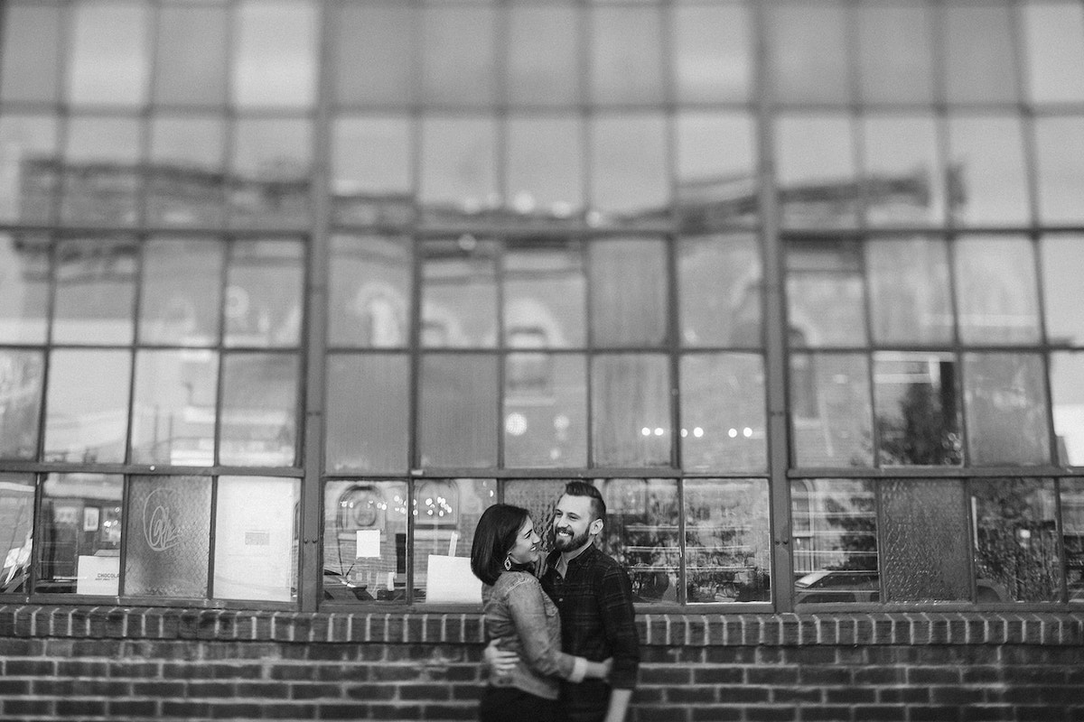 man and woman hugging in front of tall metal-framed windows and brick building for Denver RiNo engagement photos