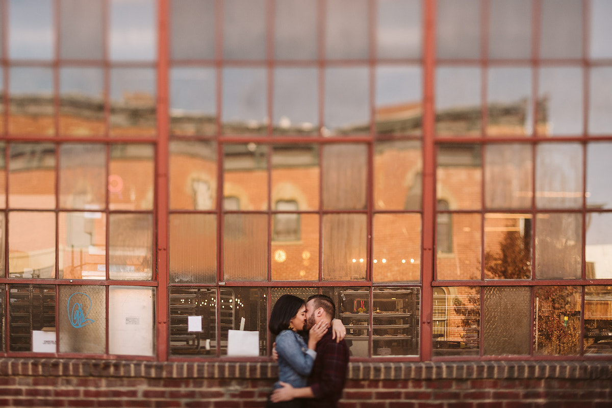 man and woman kissing in front of tall metal-framed windows and brick building for Denver RiNo engagement photos
