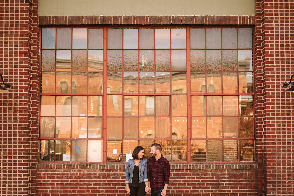 man and woman holding hands in front of tall metal-framed windows and brick building for Denver RiNo engagement photos