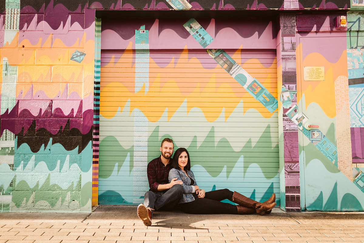 man and woman sit on the ground leaning against a brightly painted wall for Denver RiNo district engagement photos