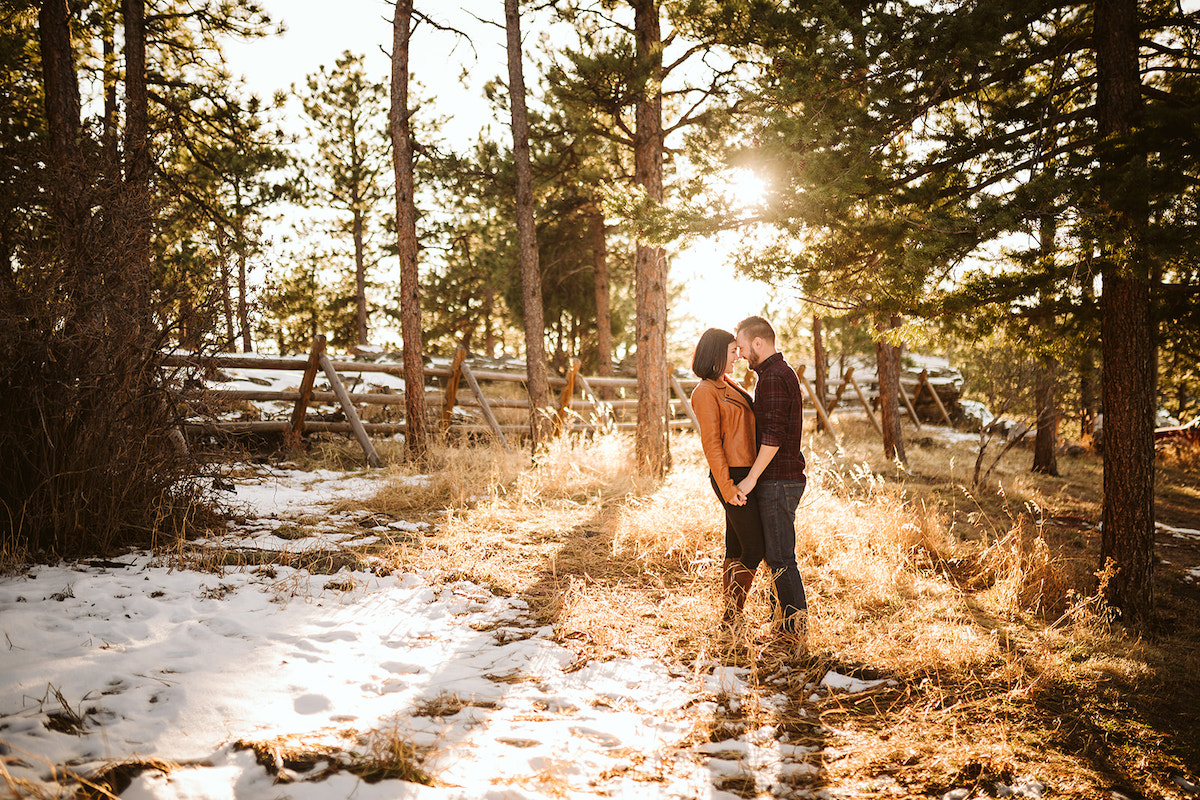 Man and woman stand in a tall grass field near snow. They hold hands and press their foreheads together as the sun sets