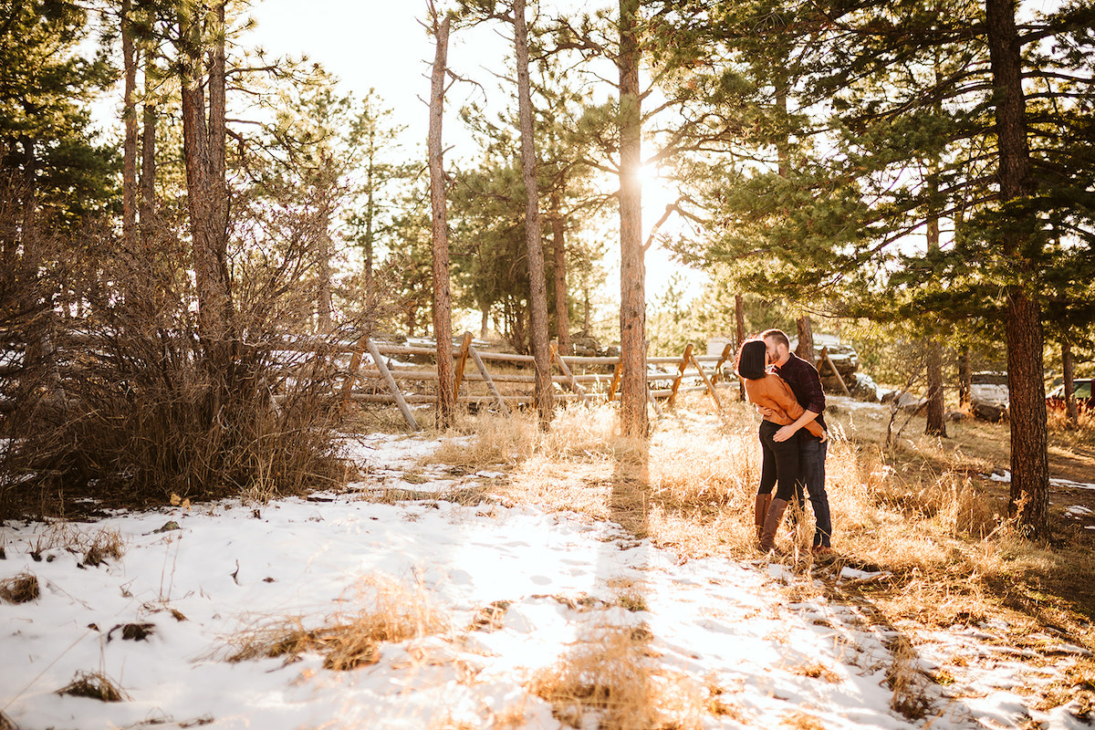 Man and woman stand in a tall grass field near snow. They hold hands and press their foreheads together as the sun sets