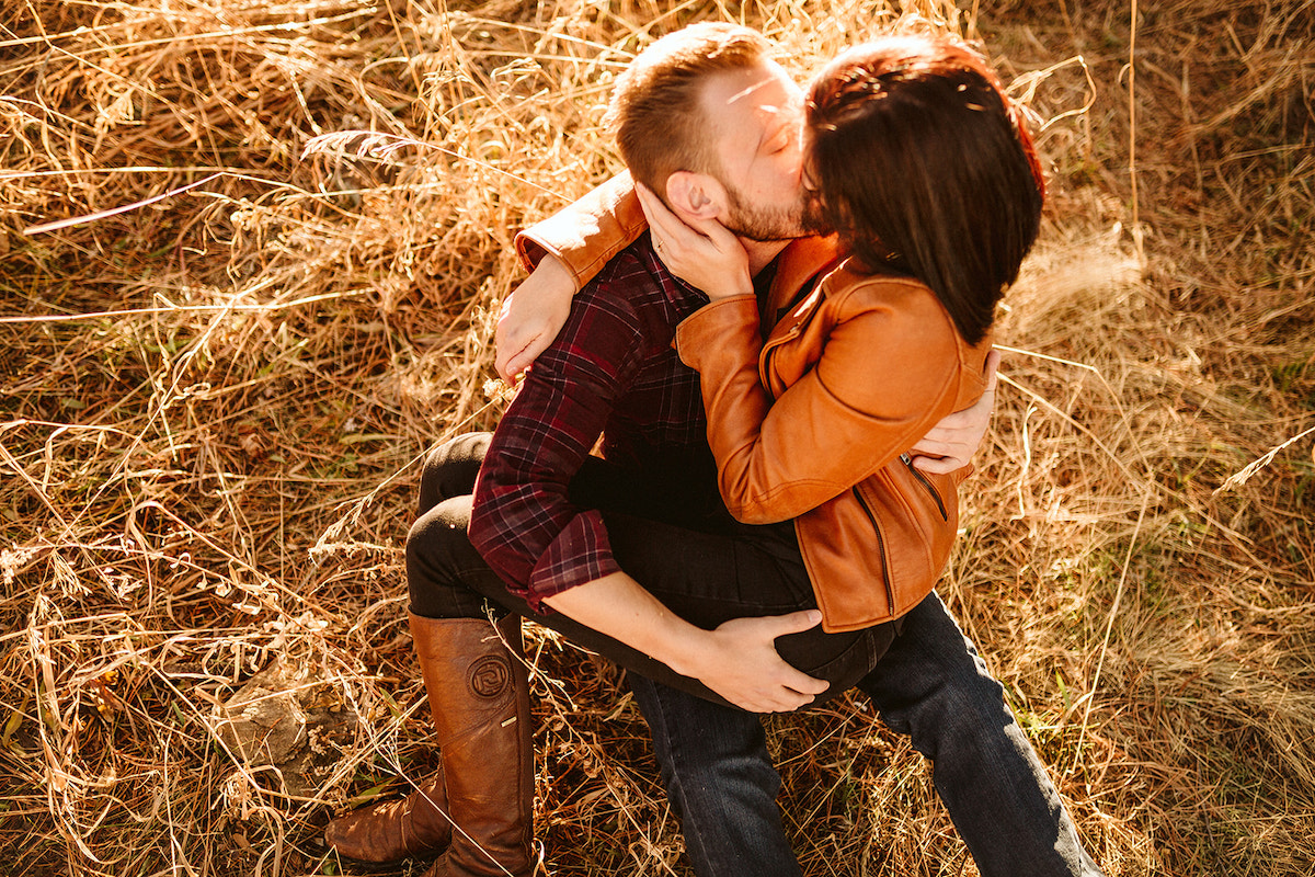 Man sits in tall golden grass with woman cuddling on his lap. She holds his cheek and kisses him.