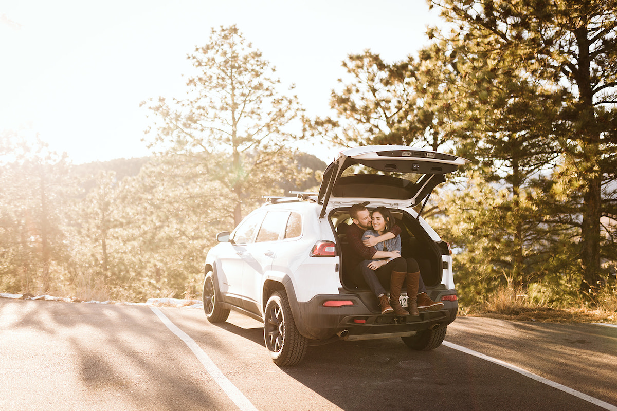 Man and woman sit in the open hatchback of their Jeep. She sits between his legs as he hugs her.