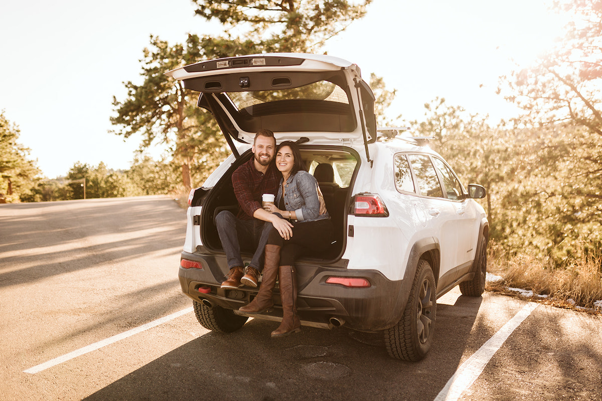 Man and woman sit in the open hatchback of their Jeep. She sits between his legs as he hugs her.