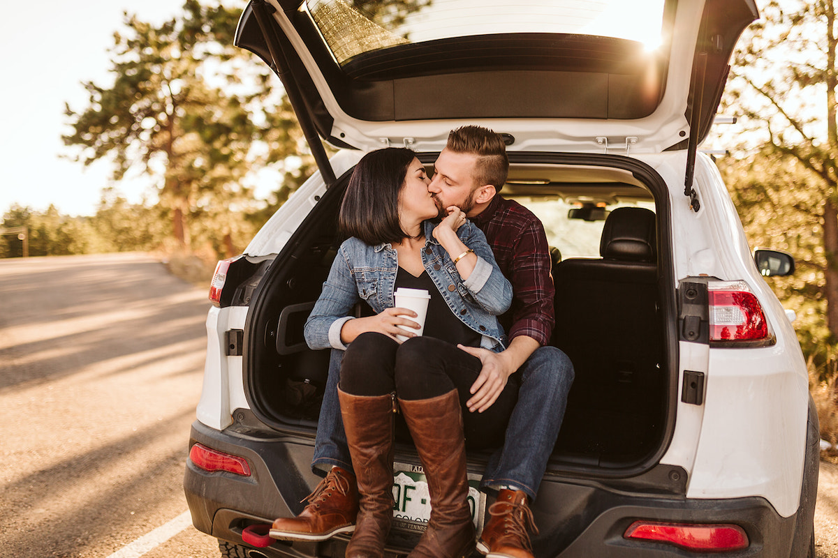 Man and woman sit in the open hatchback of their Jeep. She sits between his legs and turns her face to kiss him.