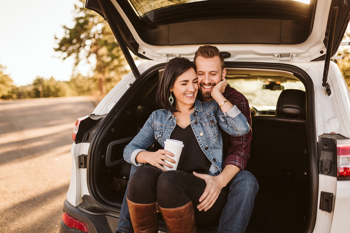 Man and woman sit in the open hatchback of white car. She sits between his legs, her hand reaching behind her to his face.