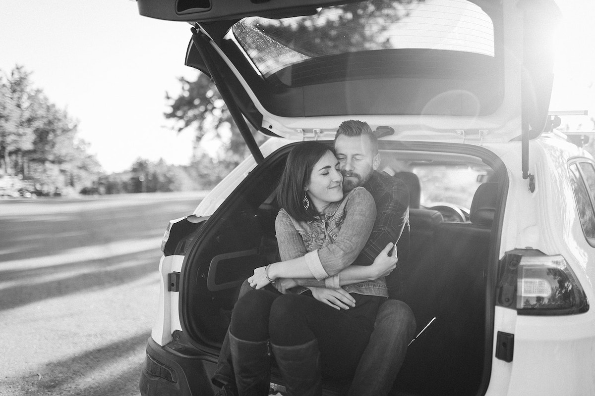Man and woman sit in the open hatchback of their white car. She sits between his legs as he hugs her.