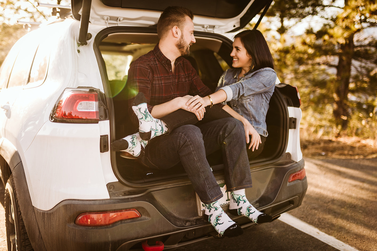 Man and woman sit in the open hatchback of white car. She has her legs over his lap. They wear white, dinosaur-printed socks