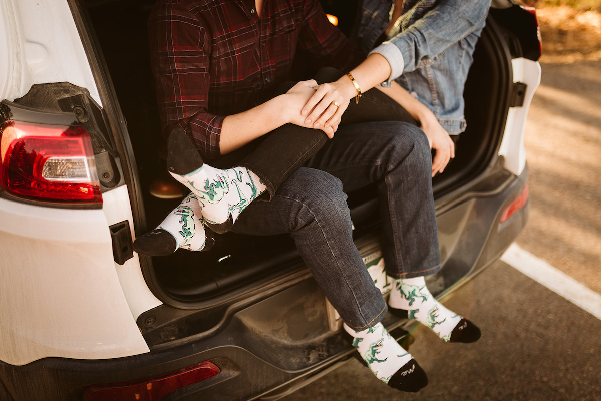 Man and woman sit in the open hatchback of white car. She has her legs over his lap. They wear white, dinosaur-printed socks