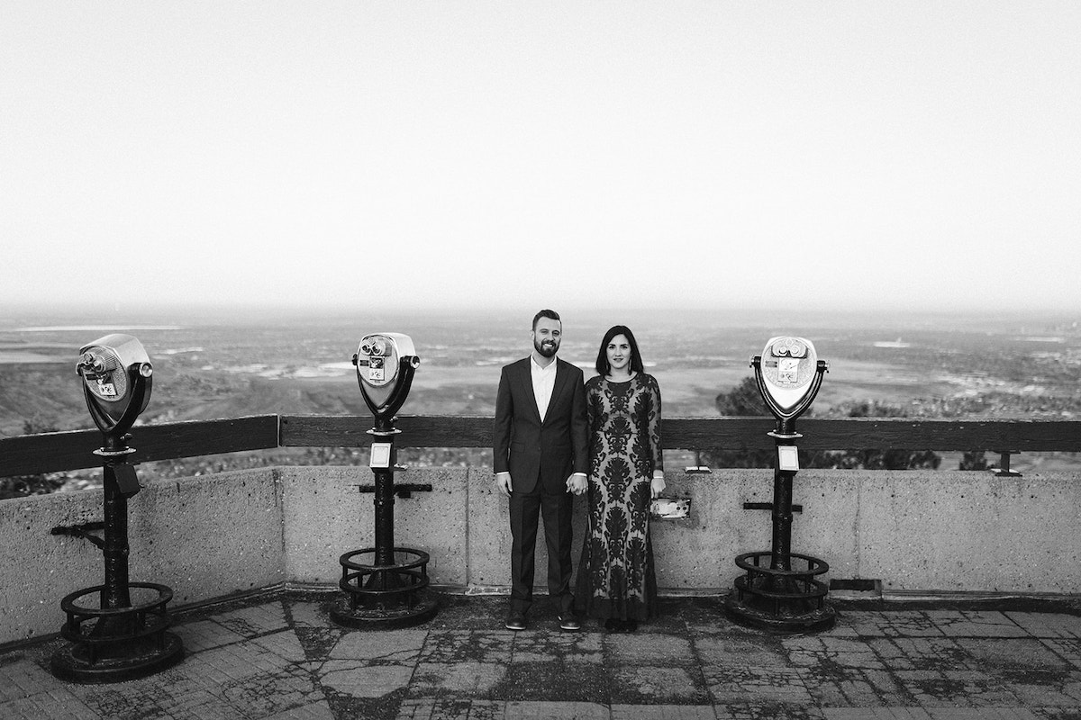 Man in dark suit hugs and woman in long maroon dress stand at an overlook between coin operated binocular machines.