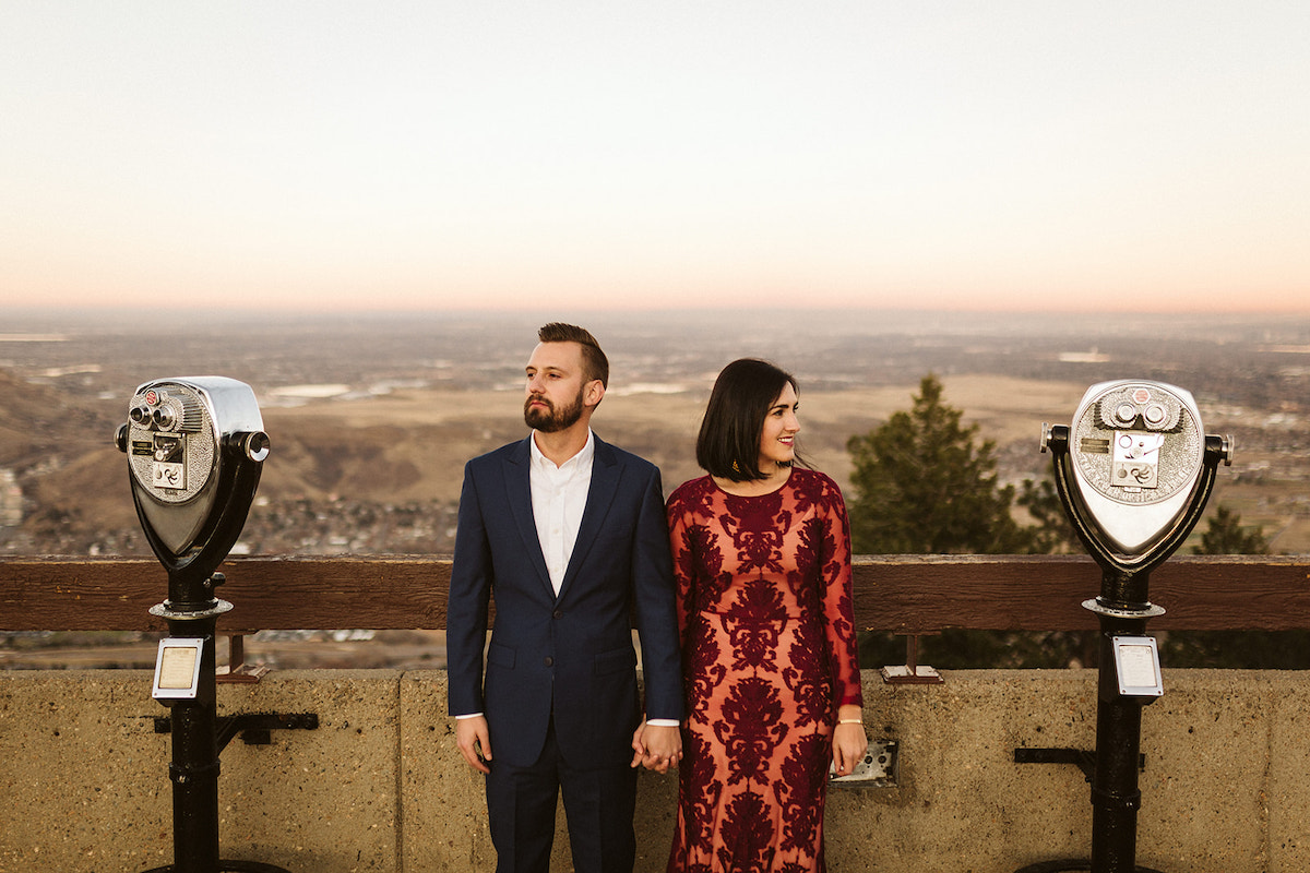 Man in dark suit hugs and woman in long maroon dress stand at an overlook between coin operated binocular machines.