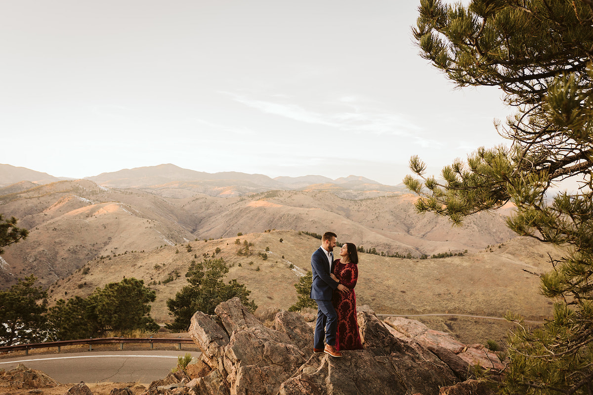 Man in dark suit and woman in long maroon dress stand on large rocks with rolling grassy mountains in the background