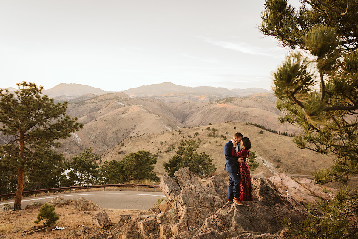 Man in dark suit and woman in long maroon dress stand on large rocks with rolling grassy mountains in the background