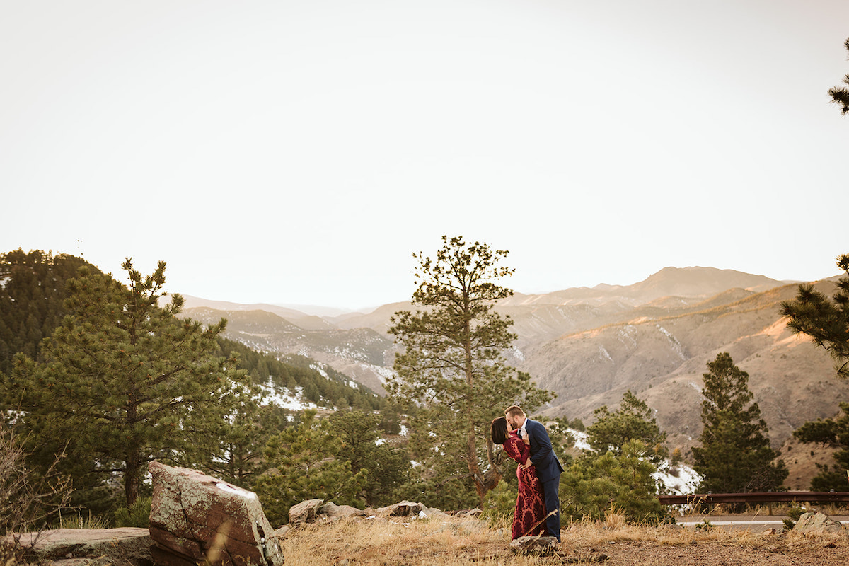 Man in dark suit and woman in maroon dress kiss in a grassy field on Colorado's Lookout Mountain for engagement photos