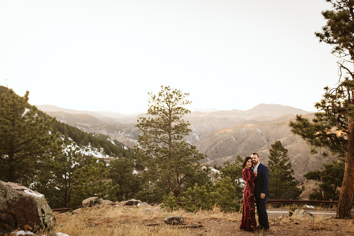 Man in dark suit and woman in maroon dress stand in a grassy field on Colorado's Lookout Mountain for engagement photos