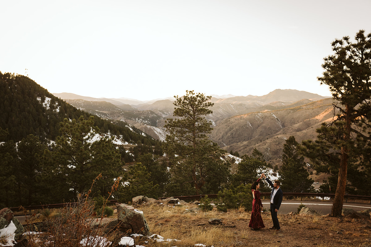 Man in dark suit twirls woman in maroon dress in a grassy field for engagement photos on Colorado's Lookout Mountain