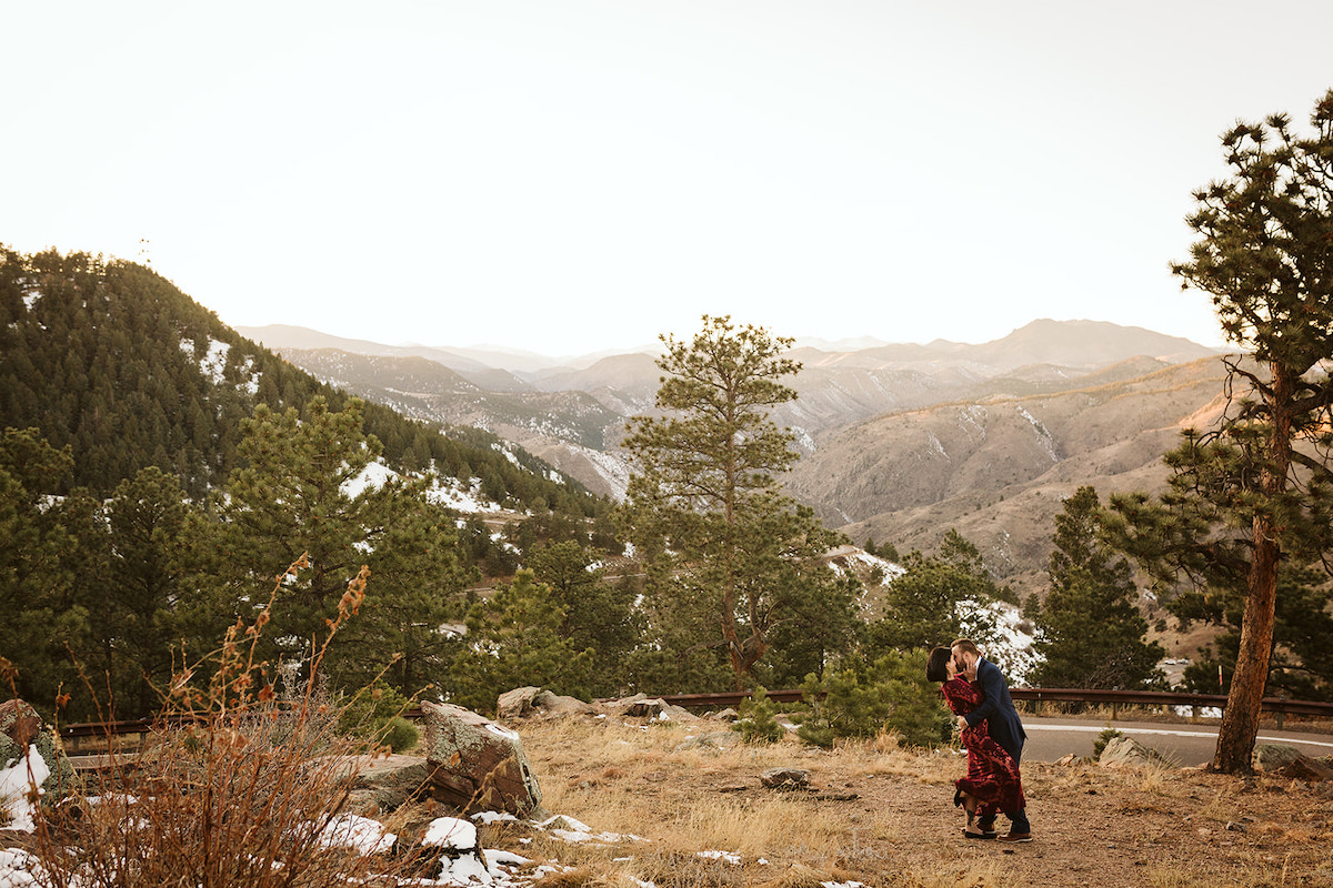 Man in dark suit and woman in maroon dress kiss in a grassy field on Colorado's Lookout Mountain for engagement photos