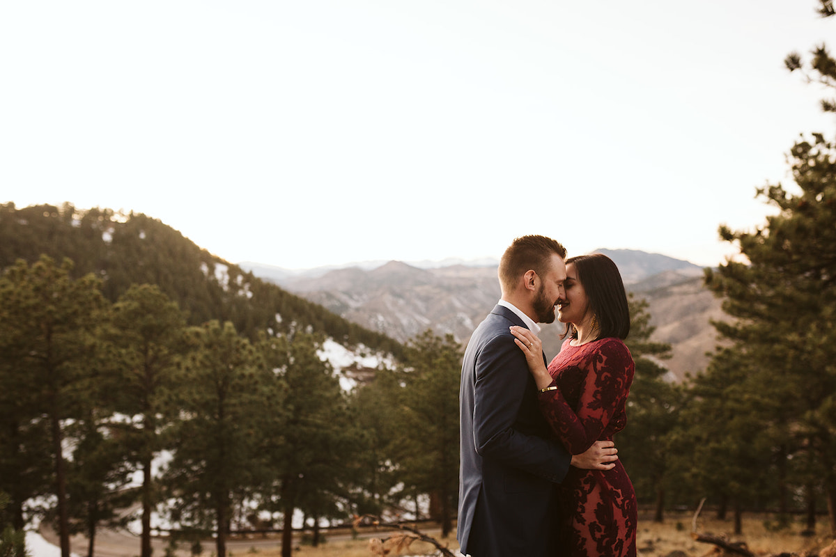 Man in dark suit and woman in maroon dress press foreheads together in front of Colorado mountain scene