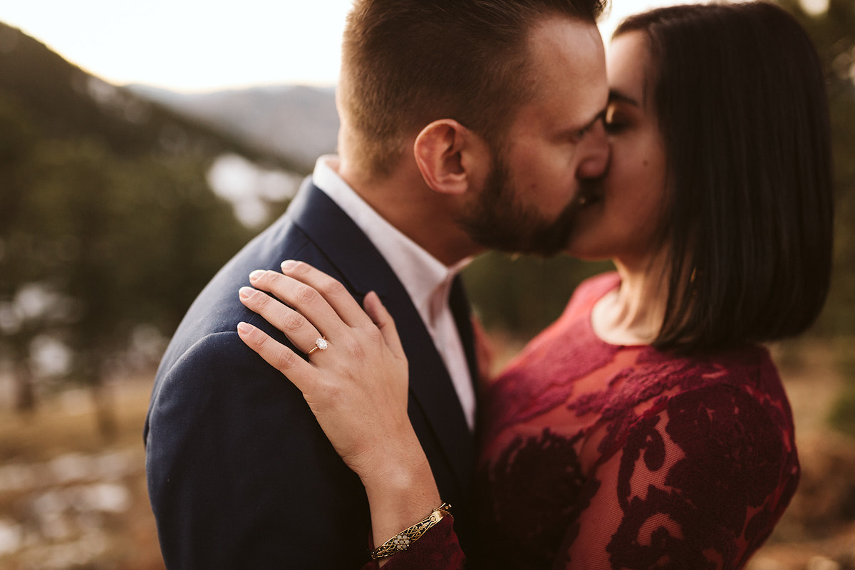 Man in dark suit and woman in maroon dress kiss, her hand and engagement ring on his shoulder