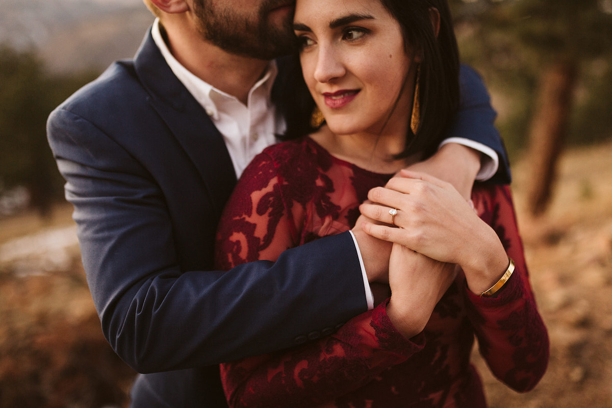 Man in dark suit wraps his arms over woman's shoulders from behind. She holds her hands over his at her chest.