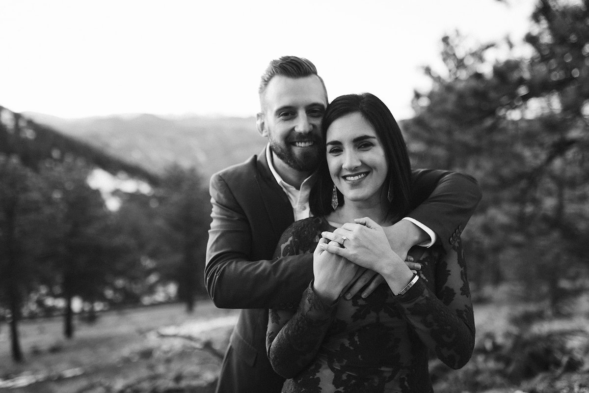 Man in dark suit hugs and woman in long maroon dress in front of Colorado mountain scenery