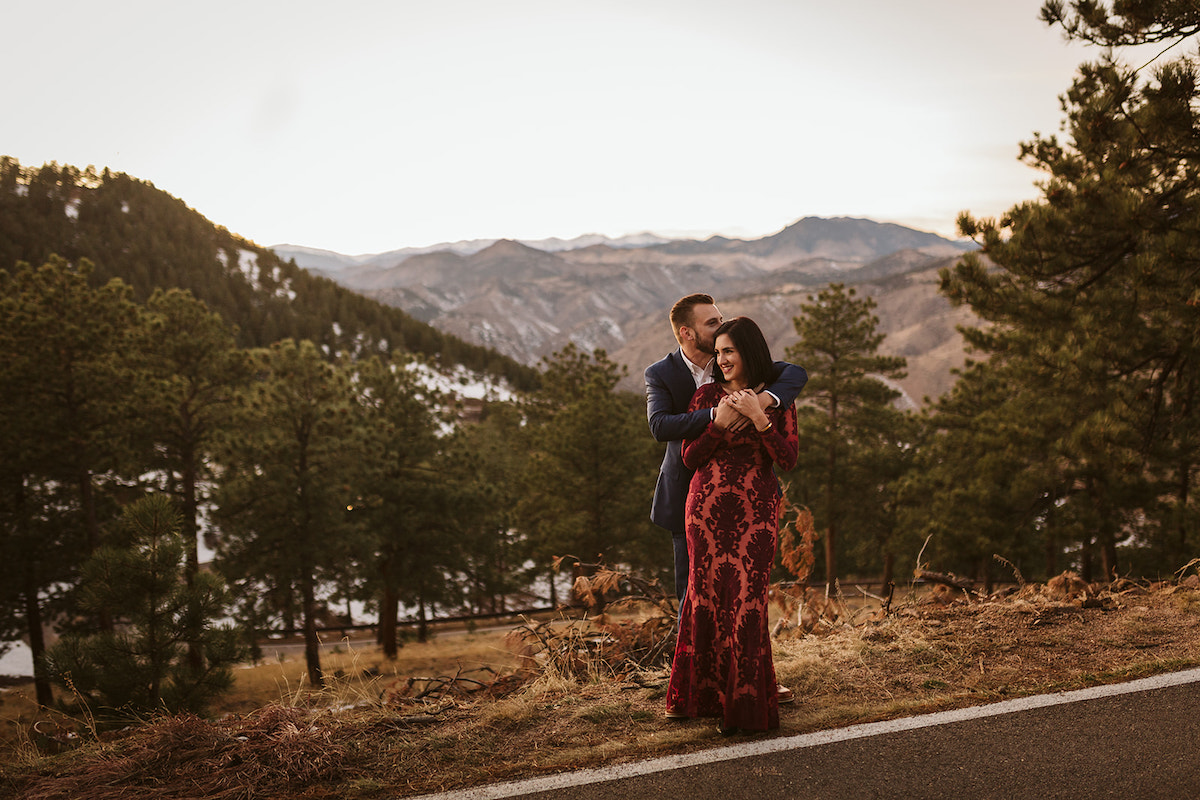 Man in dark suit hugs and woman in long maroon dress in front of Colorado mountain scenery