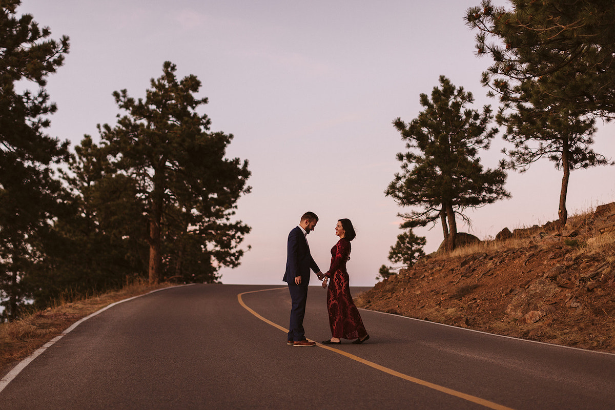 Man in dark suit and woman in long maroon dress hold hands in the middle of the street on Lookout Mountain in Colorado