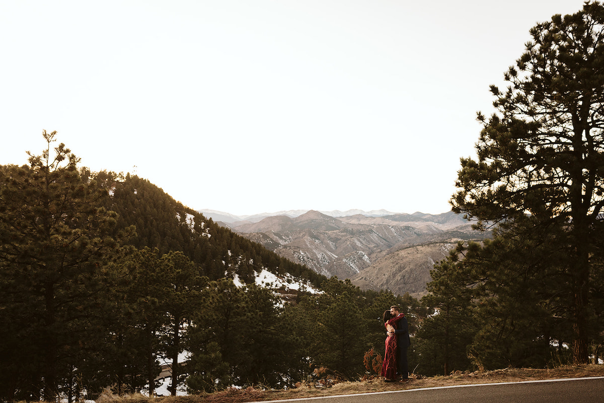 Man in dark blue suit and woman in long maroon dress hug overlooking the mountain view from Lookout Mountain in Colorado