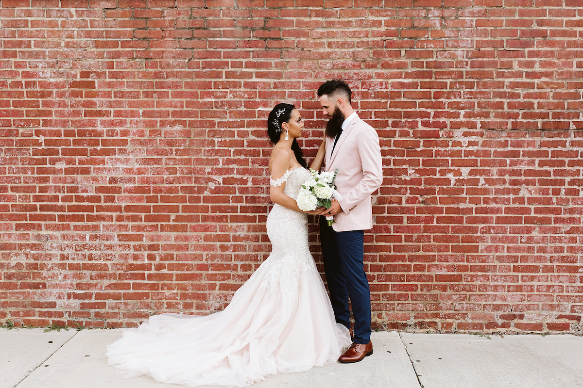 bride and groom face each other against a brick wall. her train pools behind her on the sidewalk.