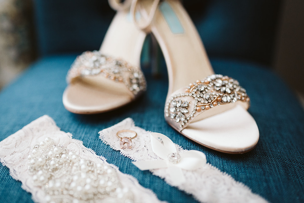 brides shoes decorated with gems sit on a rich, blue chair next to her lace garter and diamond engagement ring