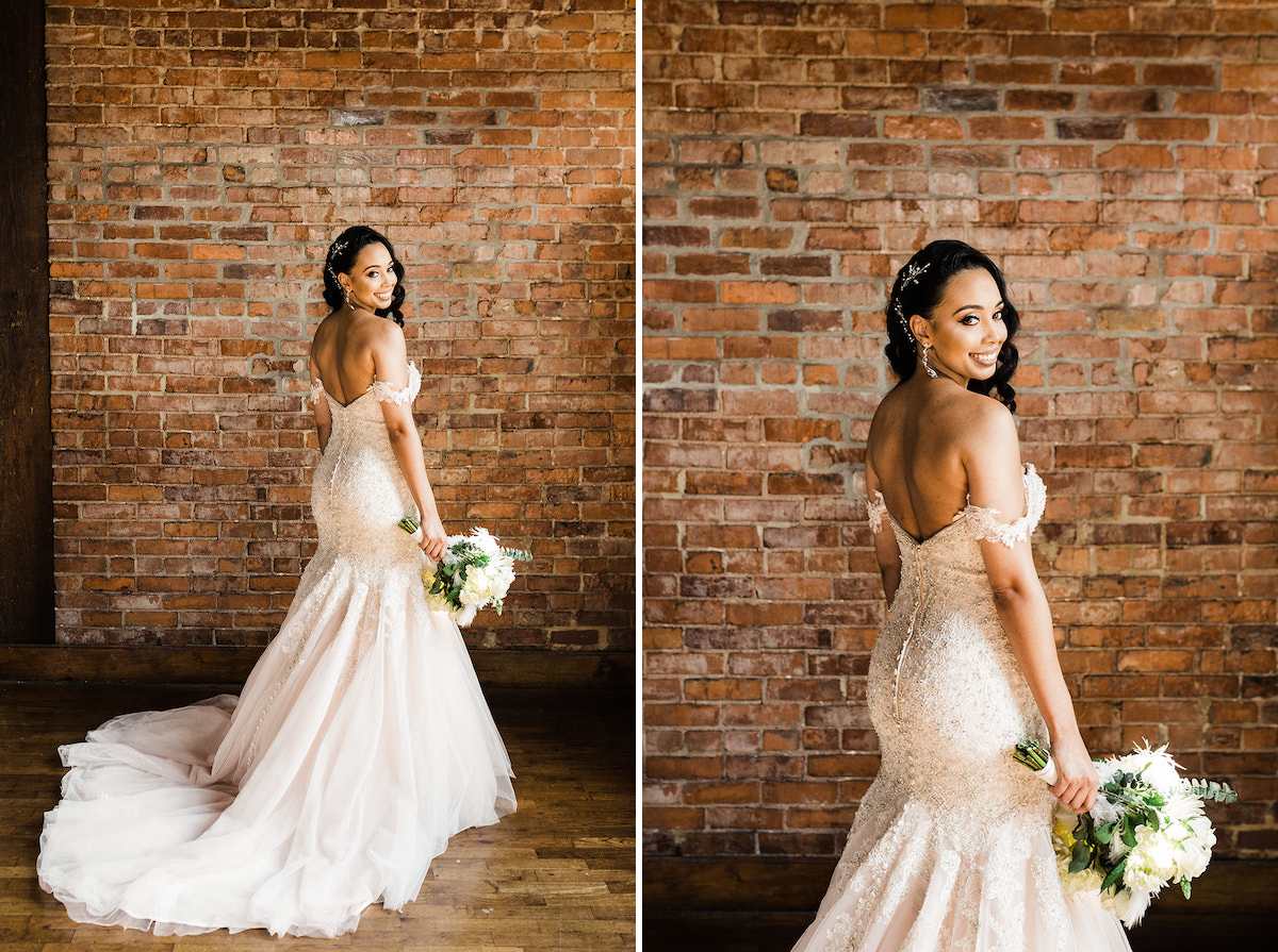 bride's long train from her mermaid wedding dress pool on wooden floor. she stands at brick wall and looks over her shoulder