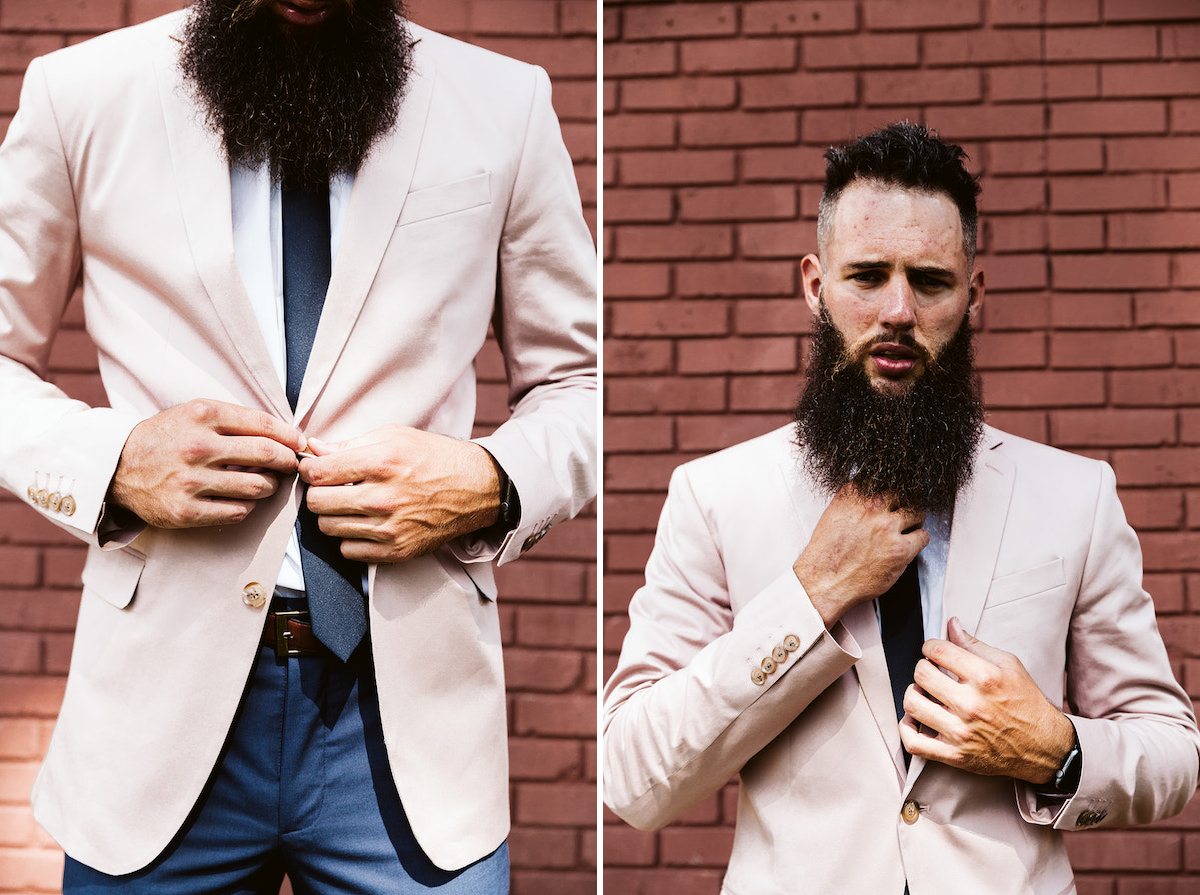 groom with long, dark beard fastens button on his pastel pink suit coat