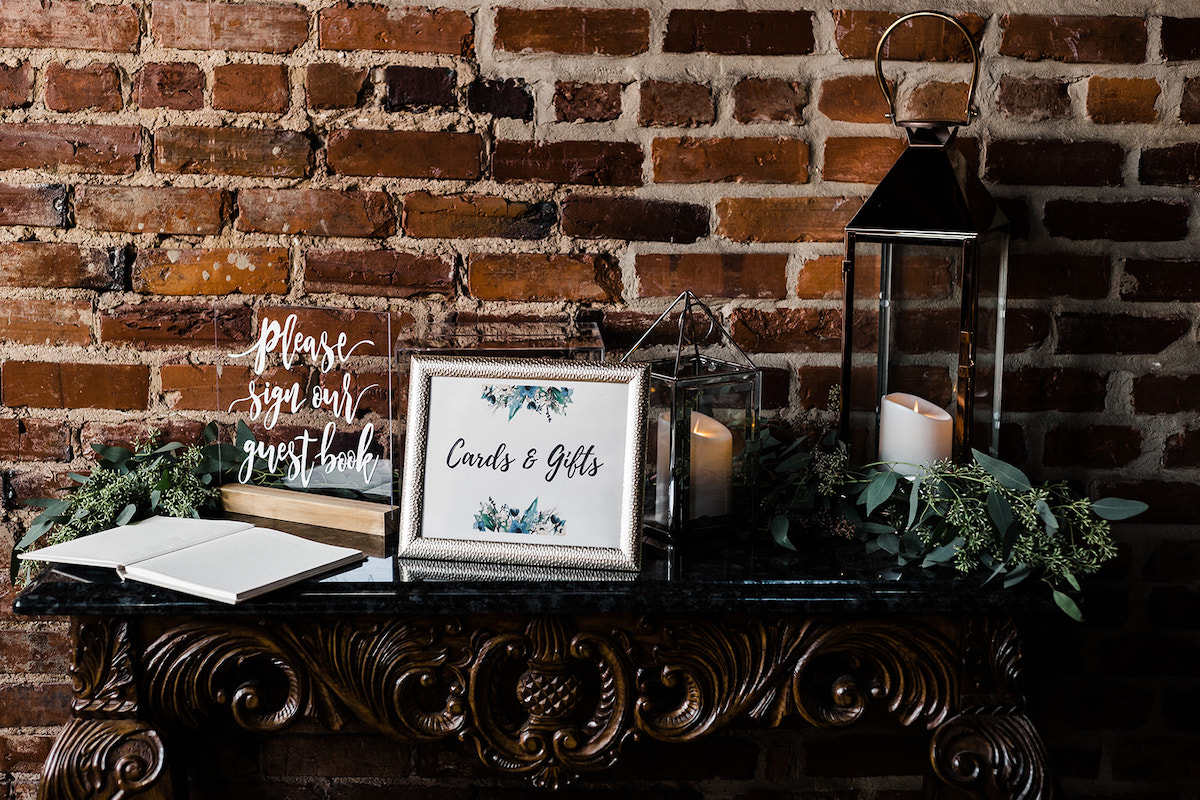 guestbook, a sign for cards and gifts, and a white candle sit on an ornate table next to a brick wall