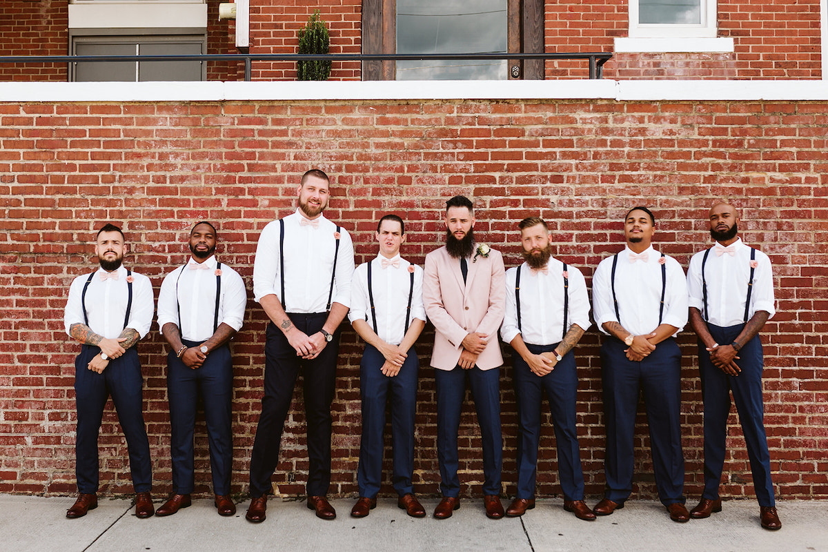 Groom and groomsmen stand in front of a tall brick wall after his Church on Main wedding