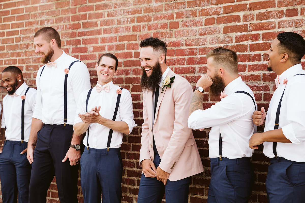 Groom and groomsmen laugh in front of a tall brick wall after his Church on Main wedding