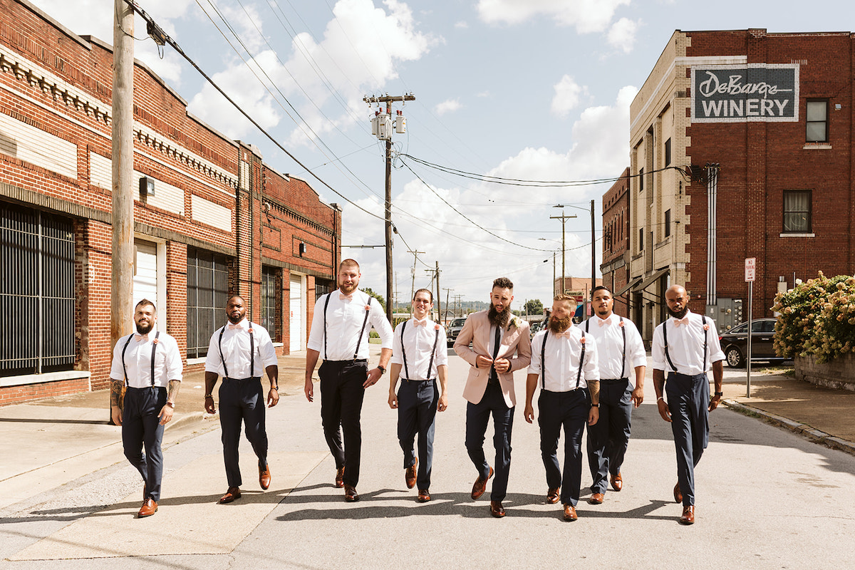 groomsmen in dark pants, white shirts, suspenders, and brown shoes walk together down the middle of the road