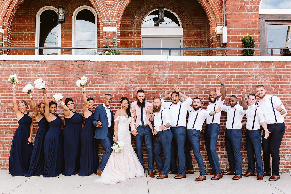 bride and groom and bridal party stand in a line in front of a tall brick wall after their Church on Main wedding