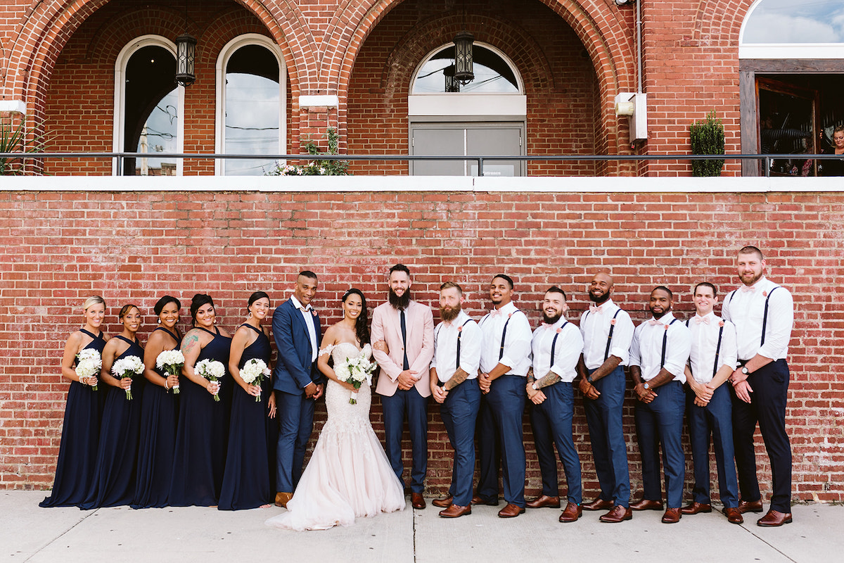 bride and groom and bridal party stand in a line in front of a tall brick wall after their Church on Main wedding