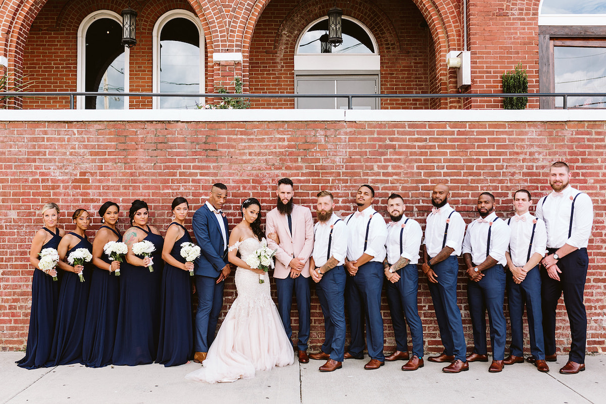 bride and groom and bridal party stand in a line in front of a tall brick wall after their Church on Main wedding