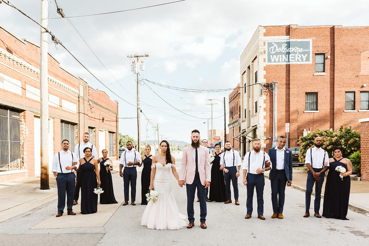 Bride and groom hold hands in the middle of the street between old brick buildings. Their wedding party stands behind them.