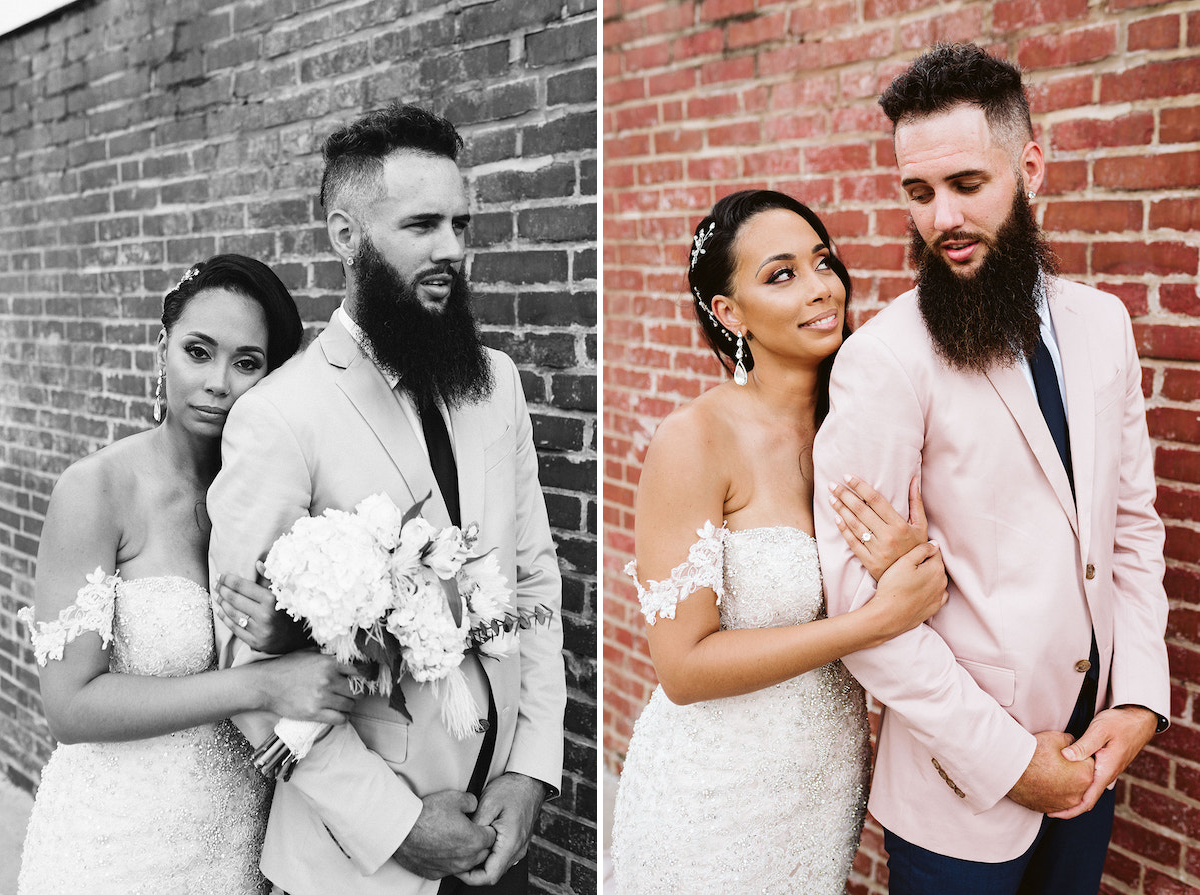 Bride stands holding groom's arm in her hands in front of a brick wall. They look at each other.