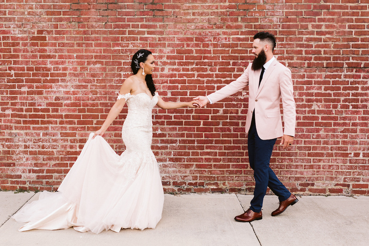 bride and groom hold hands and face each other in front of a brick wall. She holds the train of her dress in her other hand.