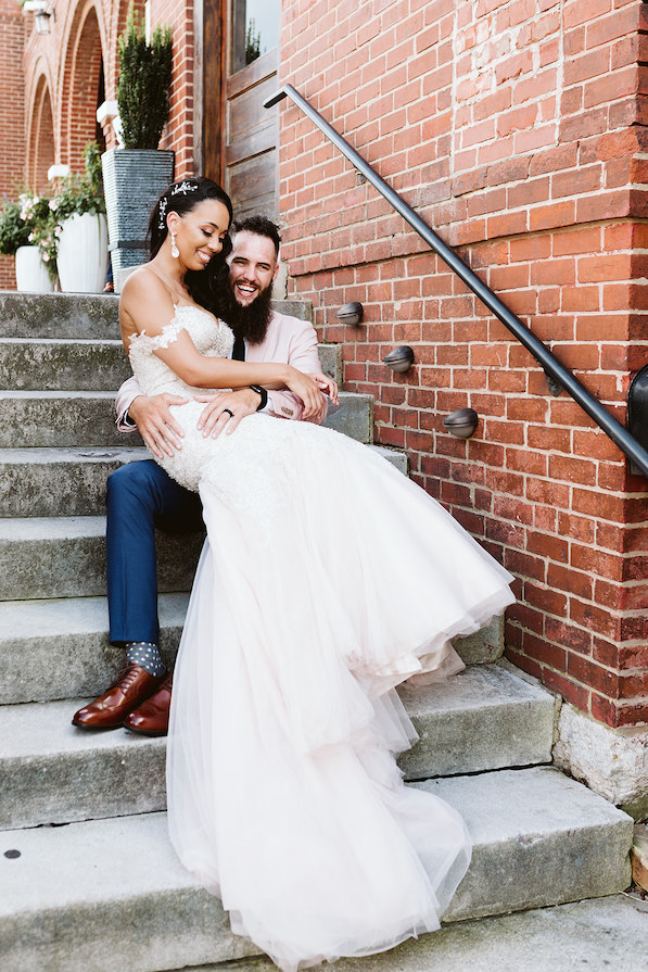 Groom sits on cement steps next to the brick exterior of The Church on Main in Chattanooga, TN. He holds bride and they laugh