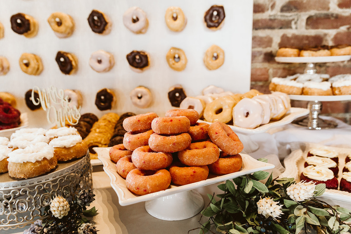 multiple flavors of donuts stacked on various plates. more donuts hang on dowels on a wall.