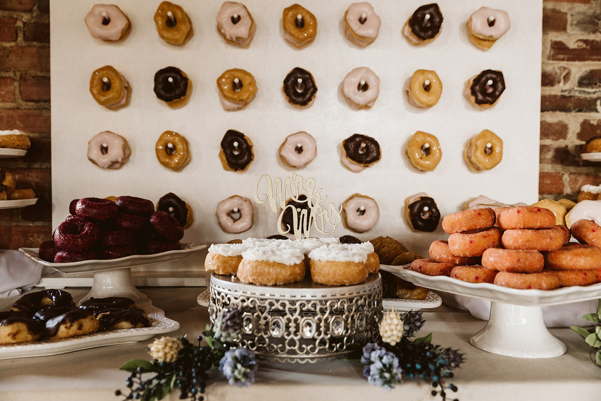 multiple flavors of donuts stacked on various plates. more donuts hang on dowels on a wall.