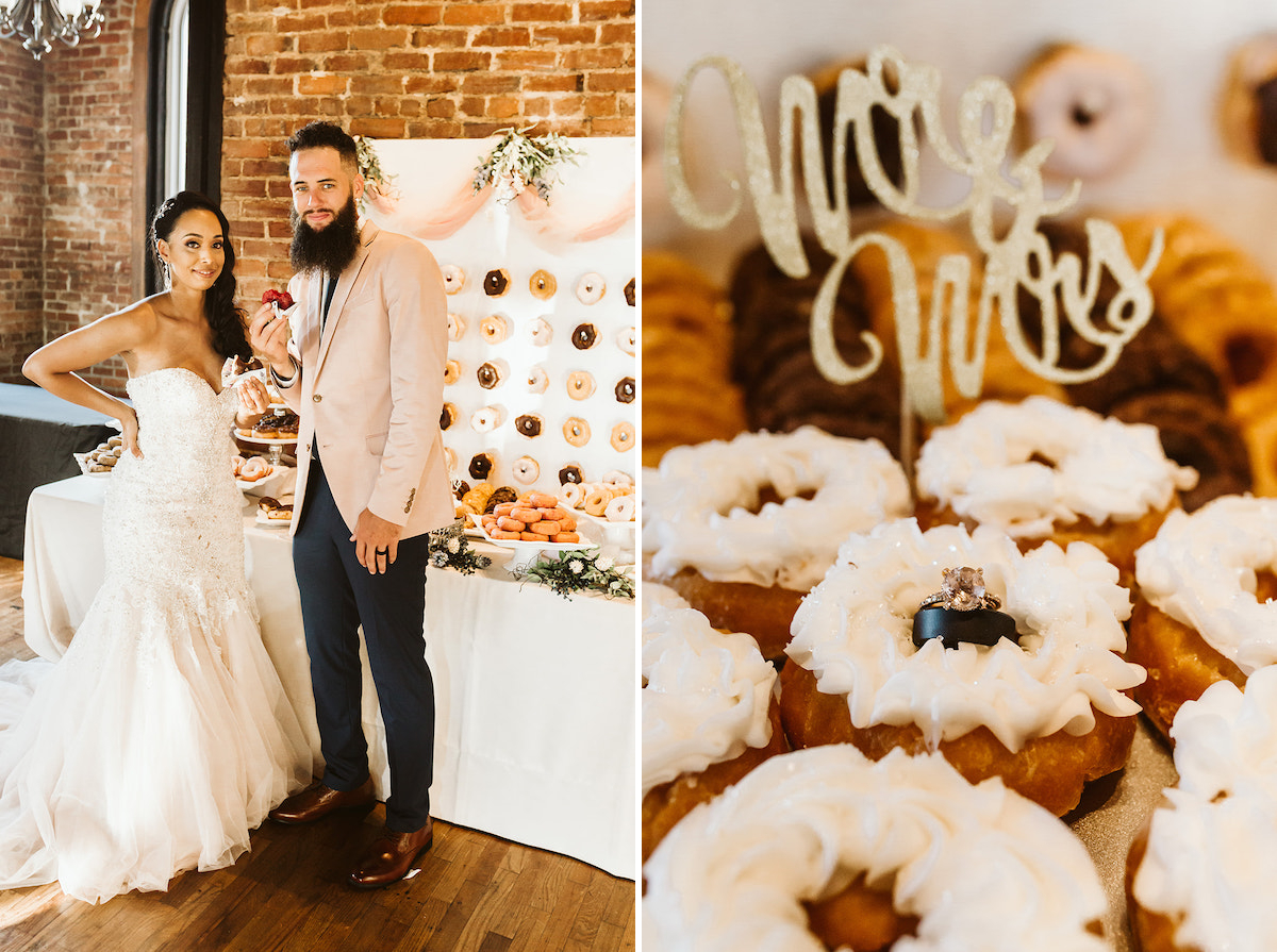 Bride and groom stand in front of a table stacked high with donuts. They each old a donut they've bitten out of.