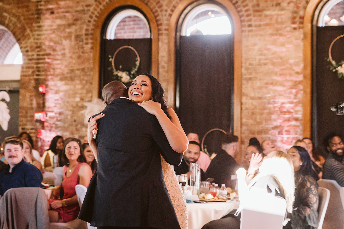 bride smiles and hugs her father after they share a dance. Tall, arched windows of The Church on Main are in the background.