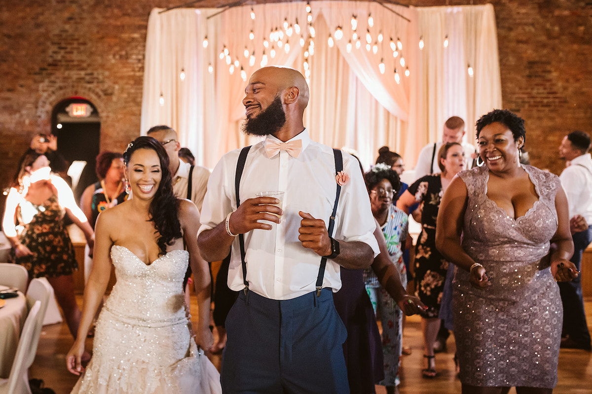 bride laughs and dances behind a tall man in white shirt and suspenders with friends around her