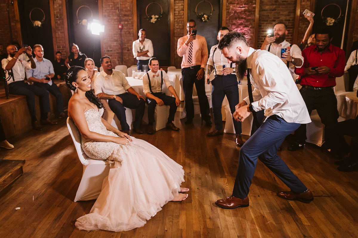 bride sits on a white chair. groom is on his knees on a wood floor in front of her as he prepares to take off her garter